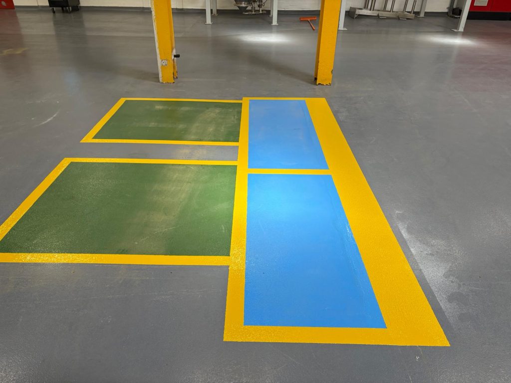 Industrial floor marking: yellow-outlined green rectangles on the left and a blue strip with yellow borders on the right, forming a safety or guide zone near columns in a workshop.