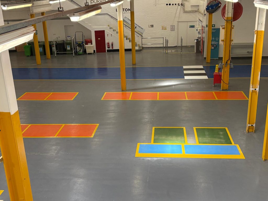 Factory floor with colored safety zones: orange hazard areas, blue walking path, and green/yellow load zones; yellow pillars and equipment in background.
