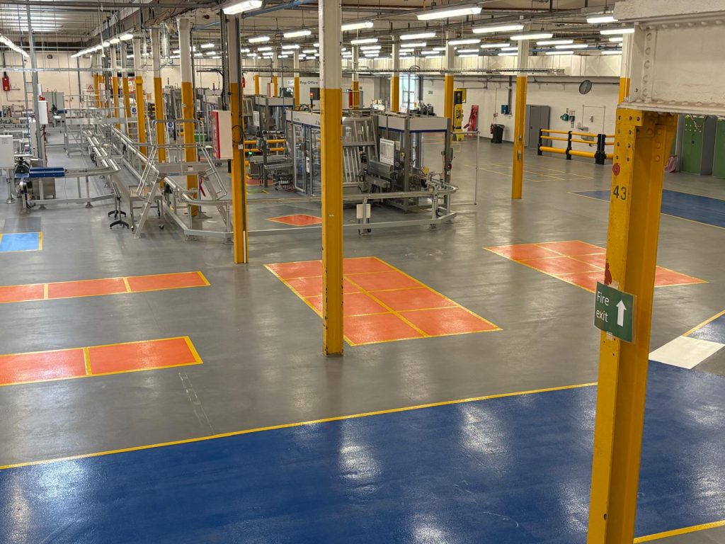 Industrial factory floor with yellow support columns, machinery, and safety zones marked in orange and red, illuminated by overhead lights.