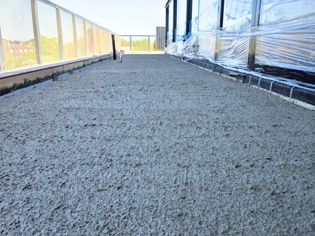 Rooftop under construction with a gravel surface, metal railing on the left, and plastic-wrapped building wall on the right.