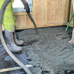 Two workers in boots spread wet concrete over a black underlayment inside a framed room, preparing the floor.