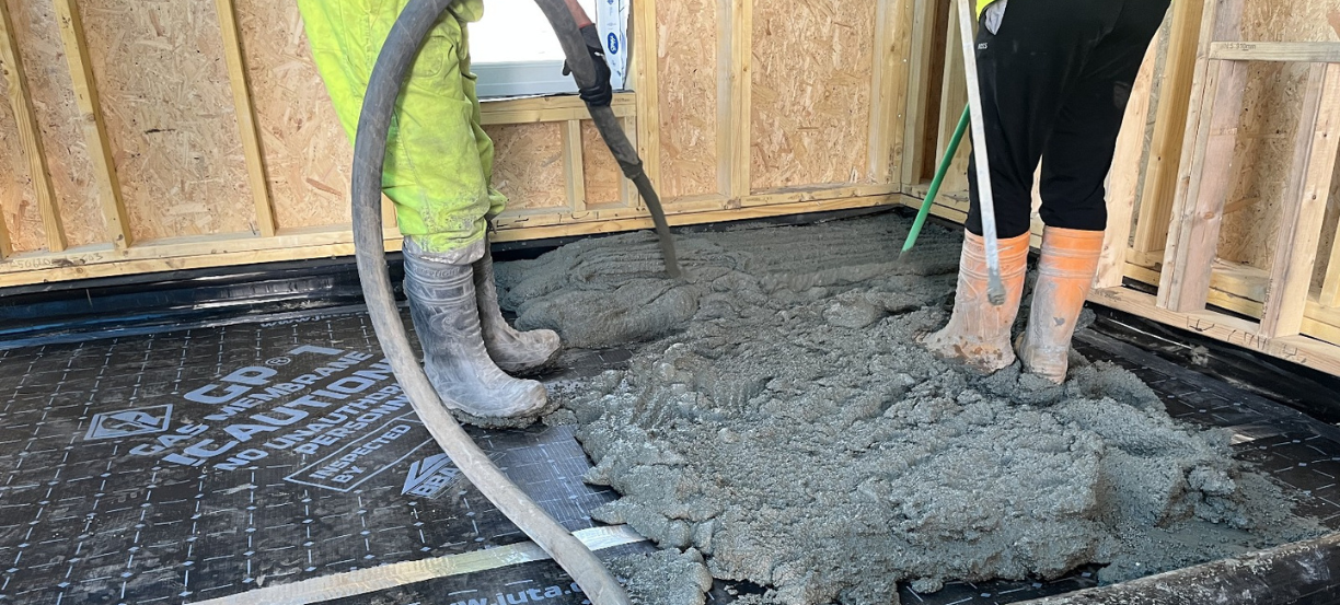 Two workers in boots spread wet concrete over a black underlayment inside a framed room, preparing the floor.