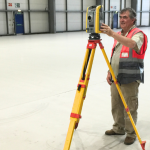 Worker in safety vest uses a surveying instrument on a yellow tripod inside a large warehouse/hangar to measure alignment.