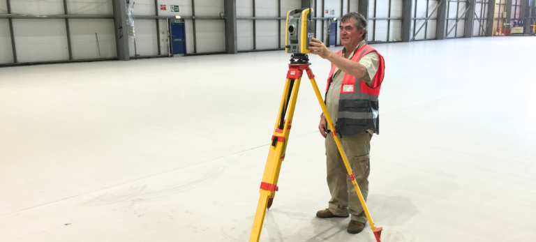 Worker in safety vest uses a surveying instrument on a yellow tripod inside a large warehouse/hangar to measure alignment.