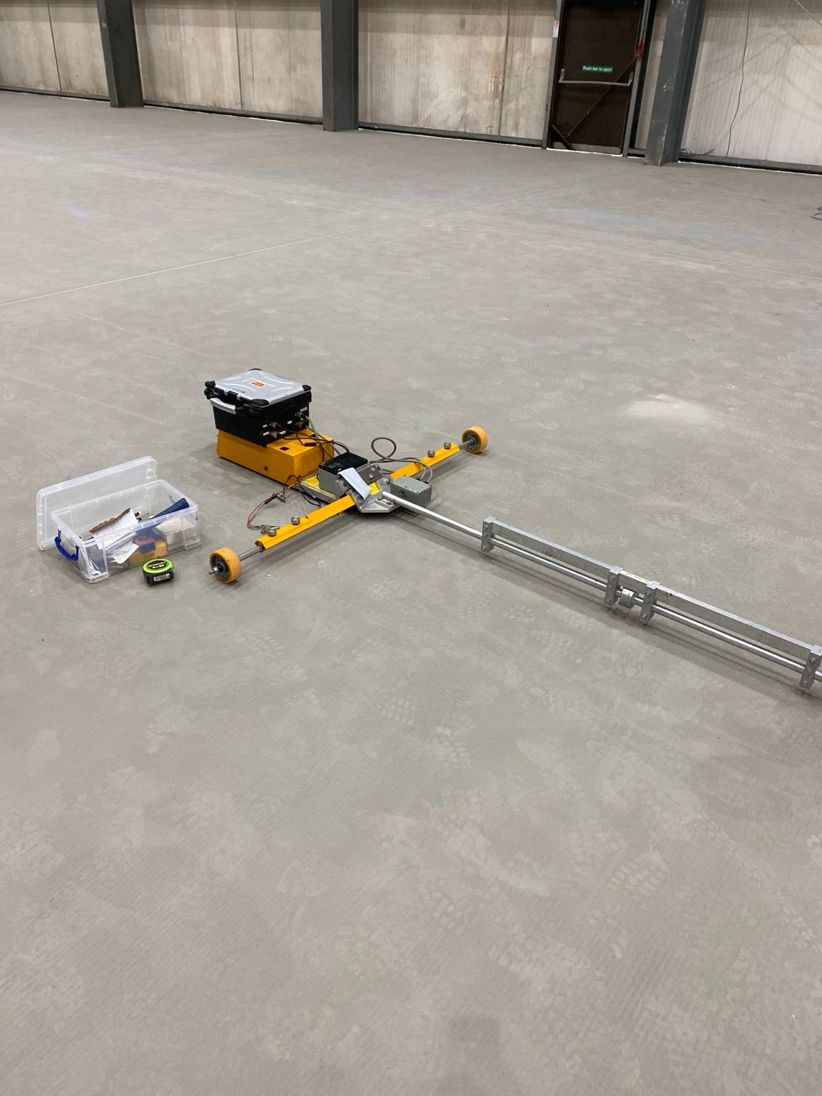 A yellow wheeled measurement trolley with a long rail on a concrete floor, nearby toolbox and gear, in a large empty space (industrial setting).