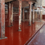Industrial plant scene with a glossy red floor, rusted metal columns, and exposed piping along a wall. Far end shows walkways and equipment.