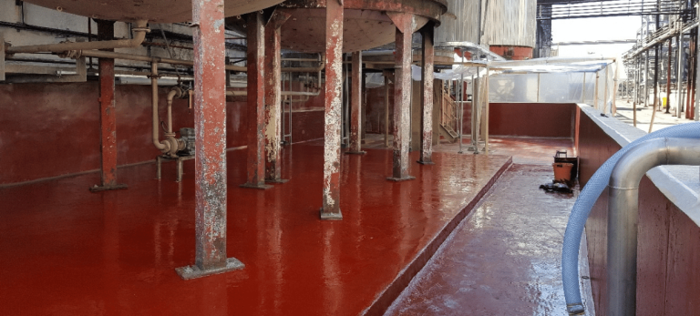 Industrial plant scene with a glossy red floor, rusted metal columns, and exposed piping along a wall. Far end shows walkways and equipment.
