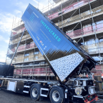 Blue dump truck unloading white gravel at a scaffolded building under construction with workers nearby in safety gear.