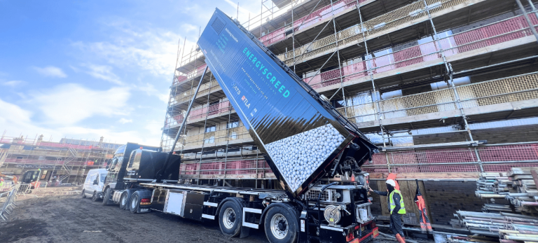 Blue dump truck unloading white gravel at a scaffolded building under construction with workers nearby in safety gear.