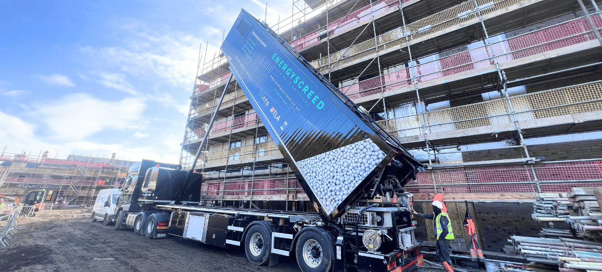 Blue dump truck unloading white gravel at a scaffolded building under construction with workers nearby in safety gear.